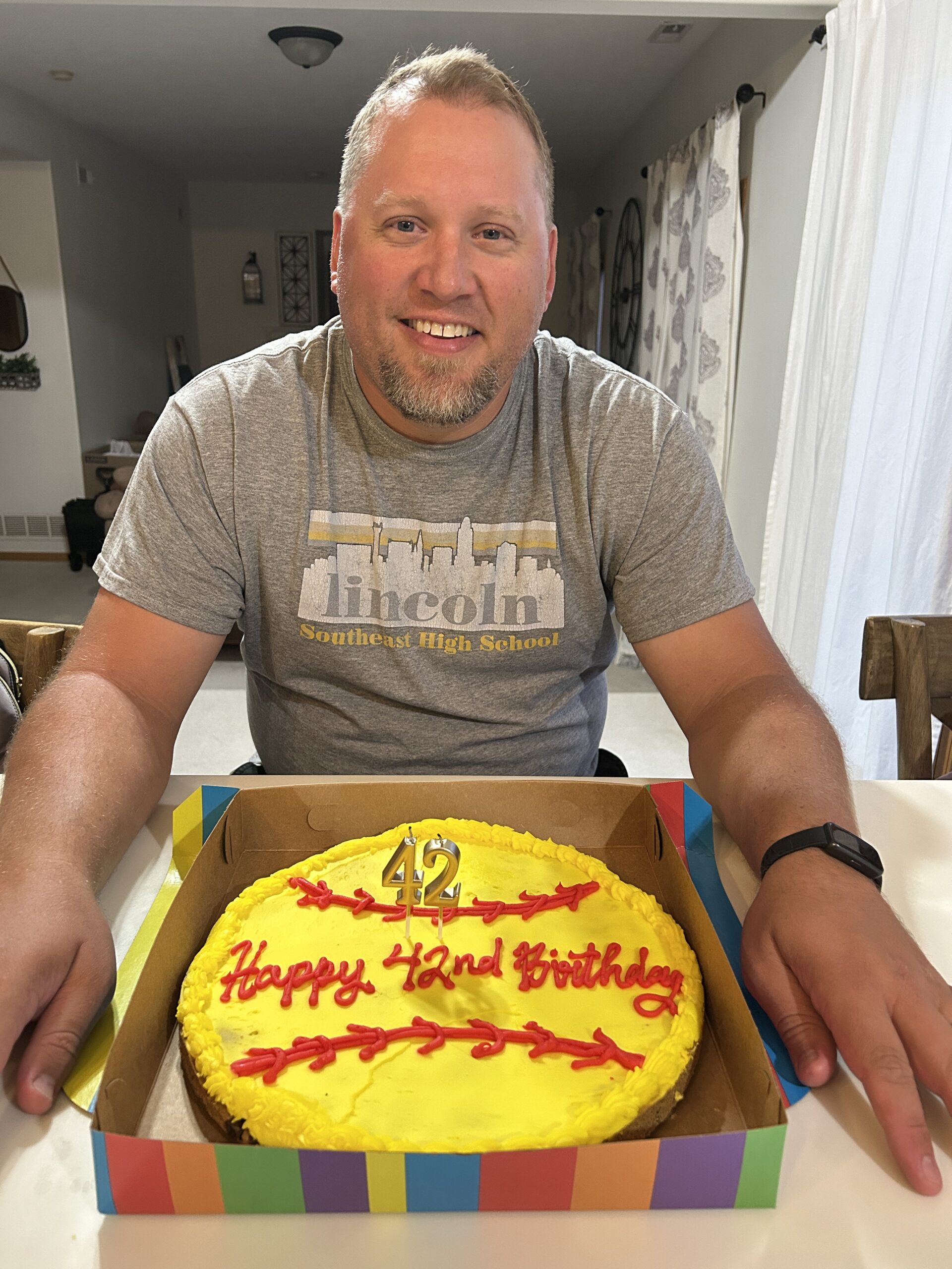 Man smiling behind a bright yellow birthday cookie cake decorated like a softball and with candles for his 42nd birthday.