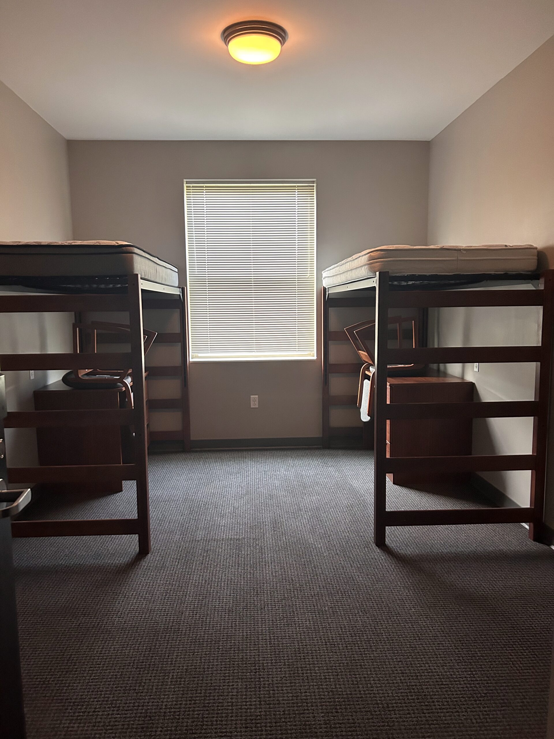 Empty college dorm bedroom with two lofted beds, desks, chairs, and a window between the beds.
