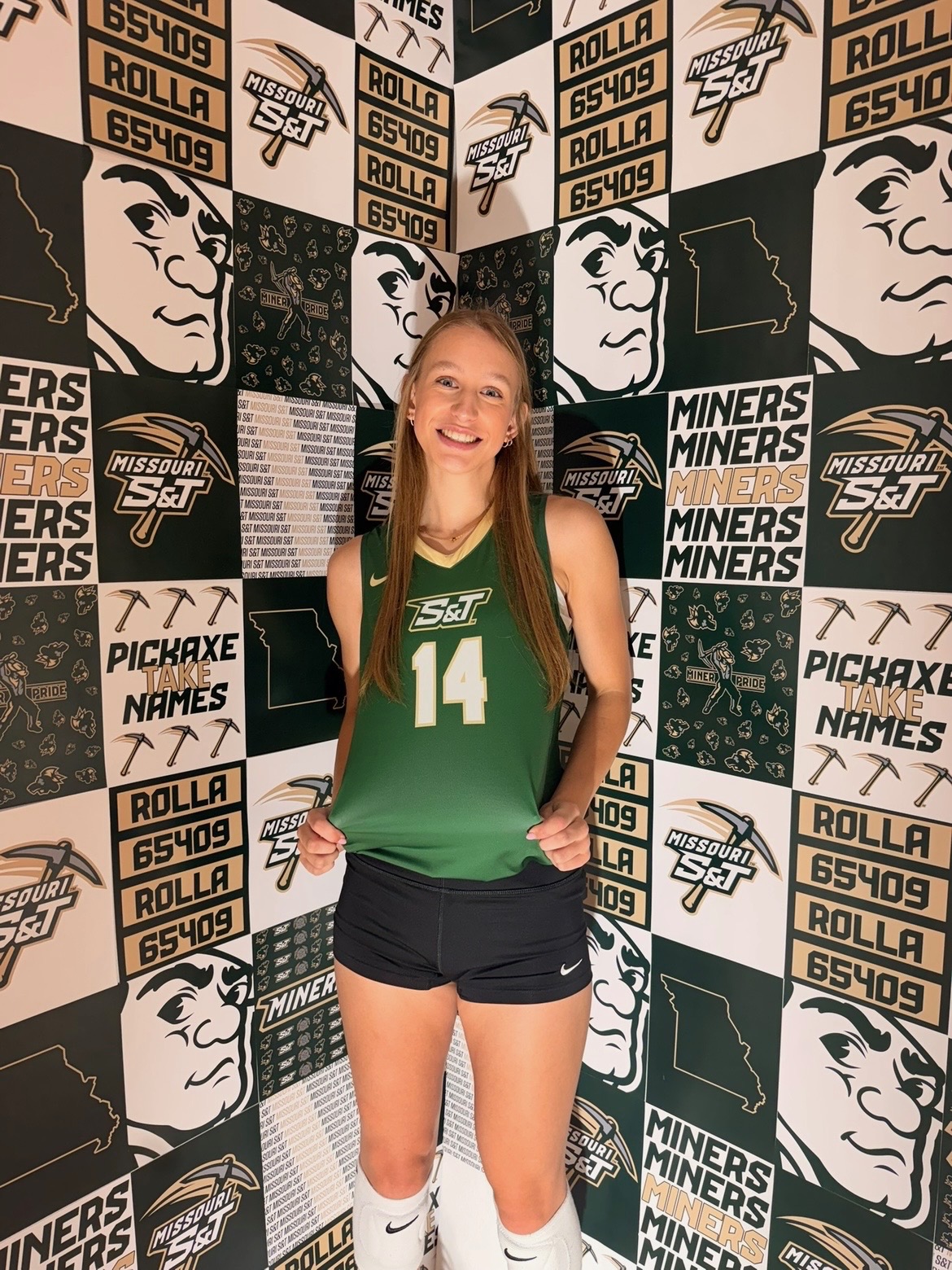 College volleyball player wearing a green Missouri S&T uniform and posing in front of a Miners media day backdrop.