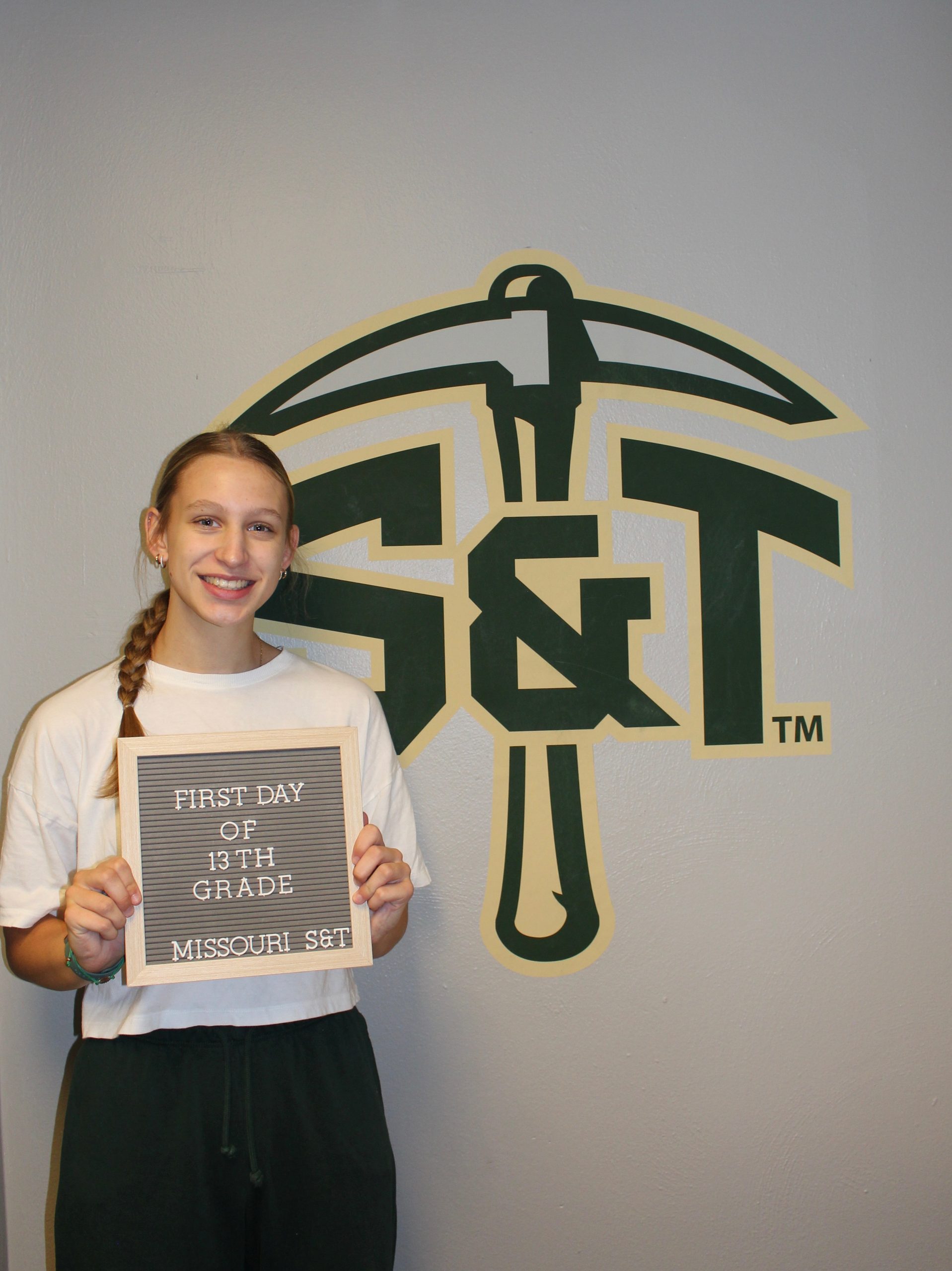 College student holding a “First Day of 13th Grade” sign in front of a large Missouri S&T Miners logo.