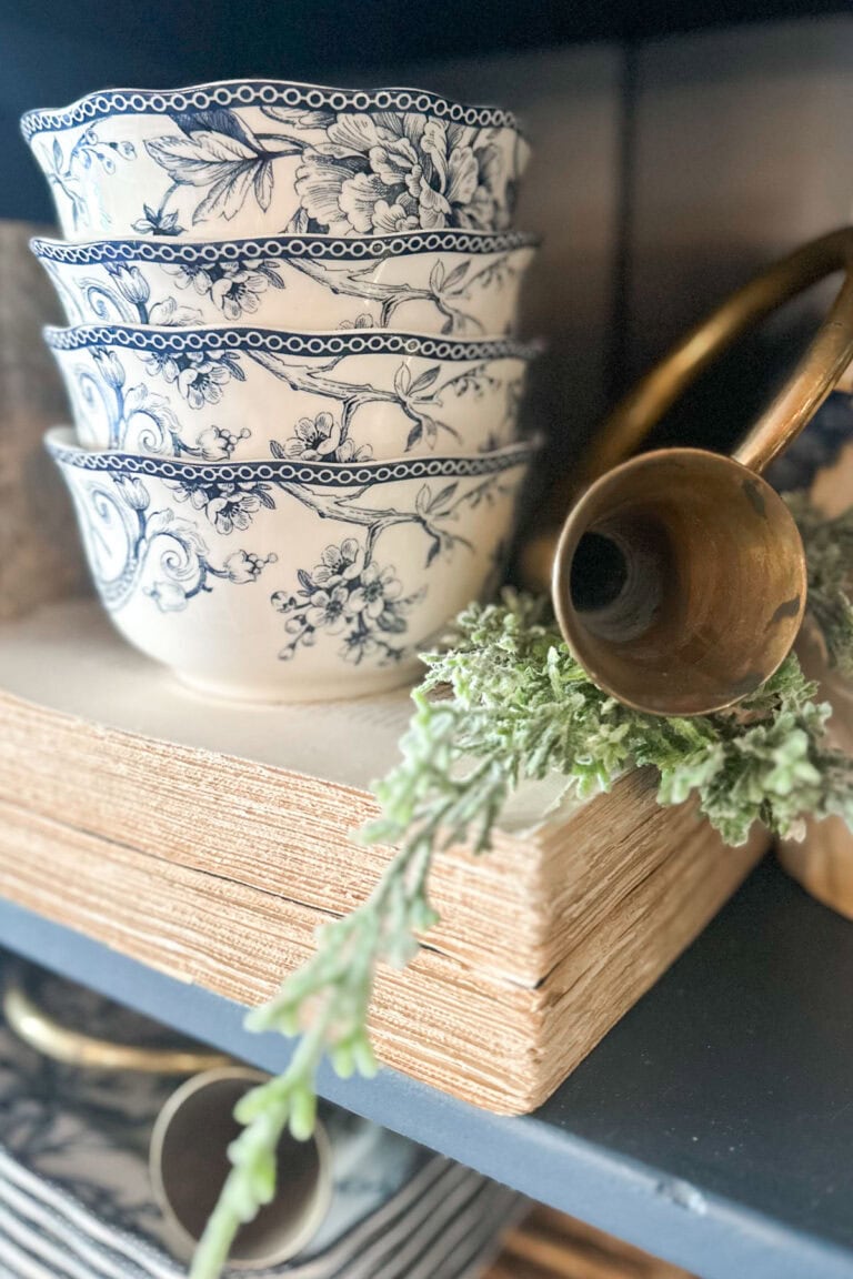 Blue and white Christmas decor with stacked floral bowls, vintage books, greenery, and a brass horn.