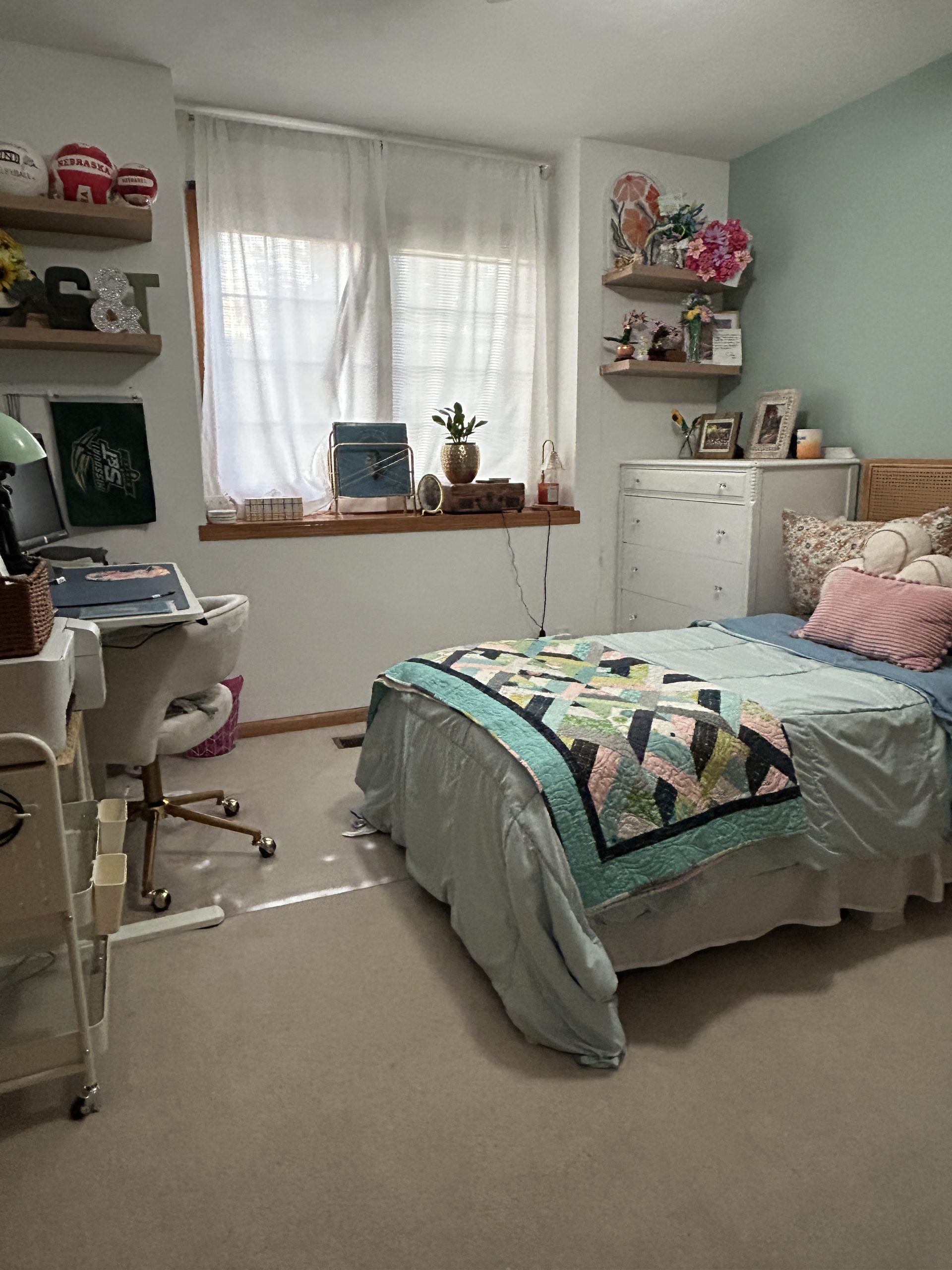 Bedroom with a bed, colorful quilt, dresser, floating shelves, and a small home office workspace by the window.