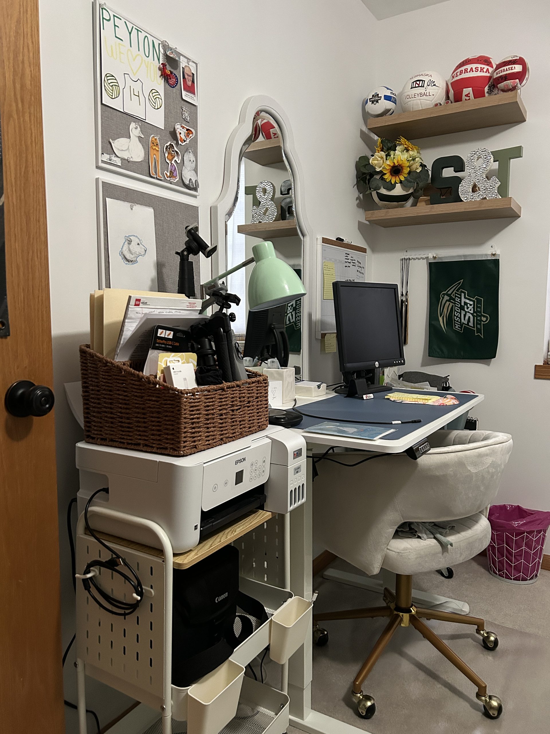 Small home office area in a bedroom with a standing desk, computer, desk chair, printer cart, floating shelves, and wall decor.