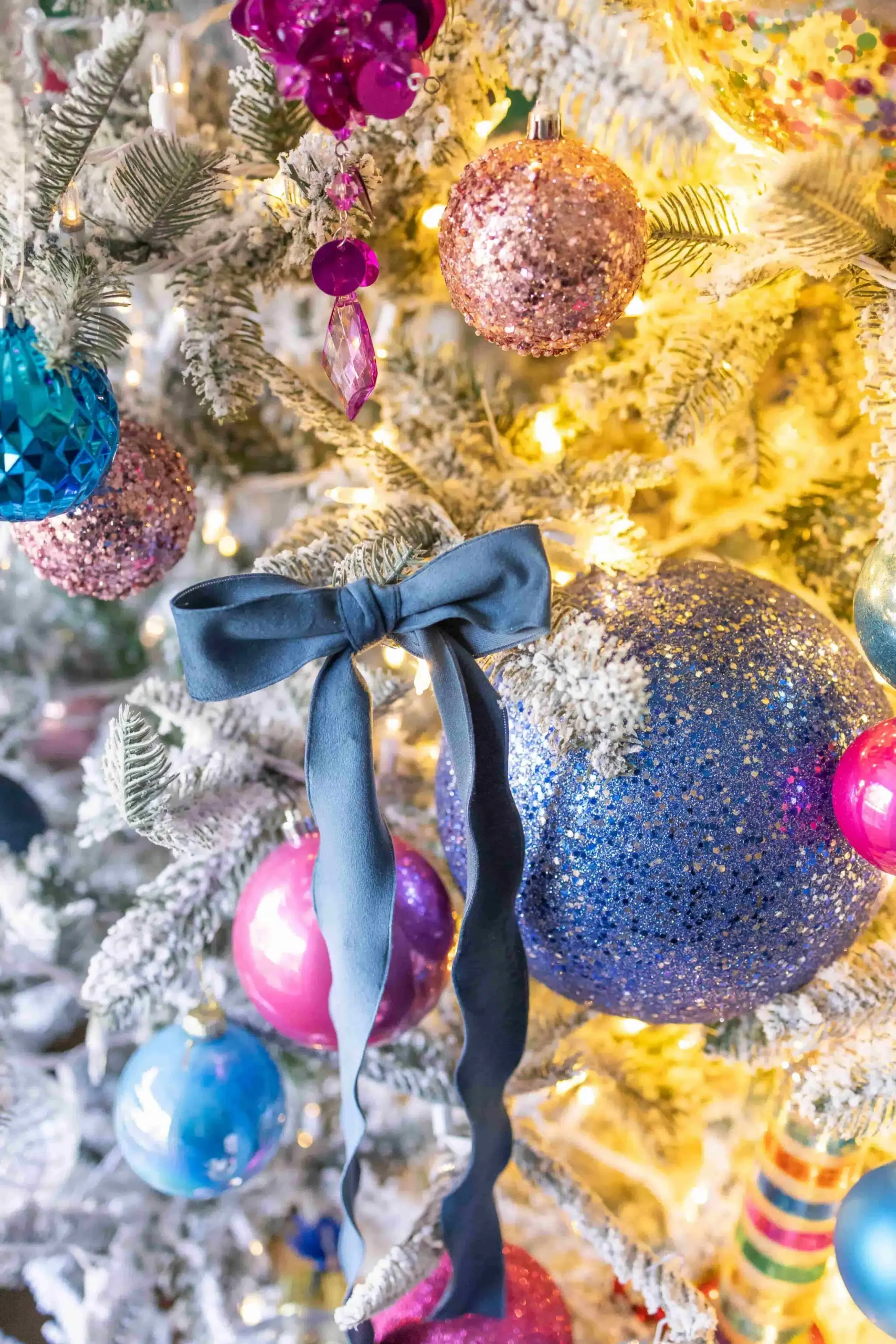 Blue and pink Christmas decor with oversized blue and pink ornaments, a blue velvet bow, and colorful accents on a flocked tree.