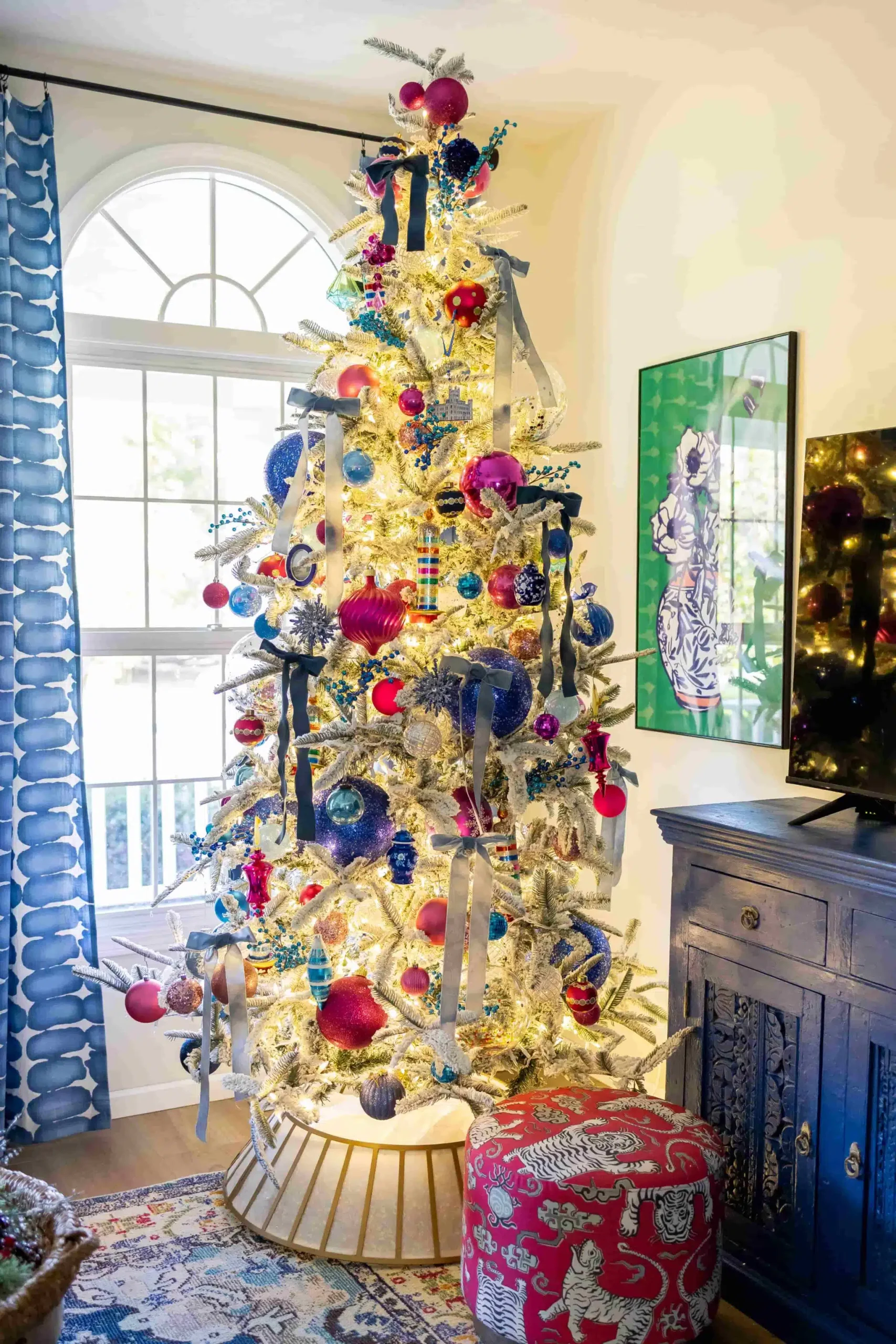 Colorful flocked Christmas tree decorated with bright blue and pink ornaments, velvet ribbon, snowflakes, and twinkle lights.