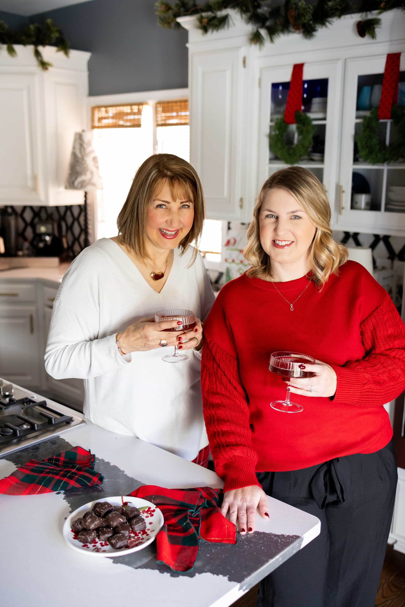 Missy Noonan and Jen Svendsen, authors of the holiday entertaining guide Twinkling Light and Festive Bites holding holiday cocktails in a Christmas-decorated kitchen during their Twinkling Lights & Festive Bites holiday gathering.