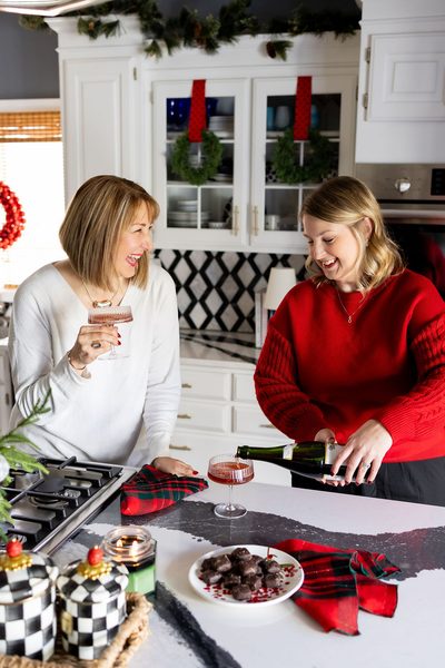 Missy and Jen laughing in a decorated Christmas kitchen as Jen pours a festive drink during a holiday gathering.