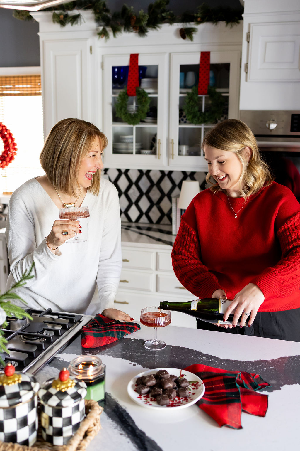 Missy and Jen laughing in a decorated Christmas kitchen as Jen pours a festive drink during a holiday gathering.