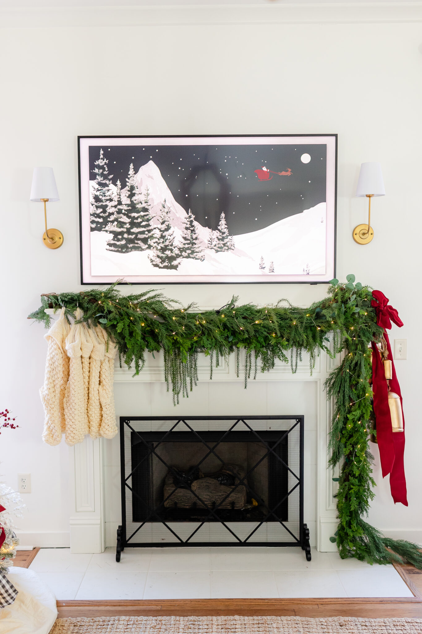Christmas fireplace mantel decorated with fresh greenery, twinkle lights, red velvet ribbon, gold bells, and cream knit stockings.