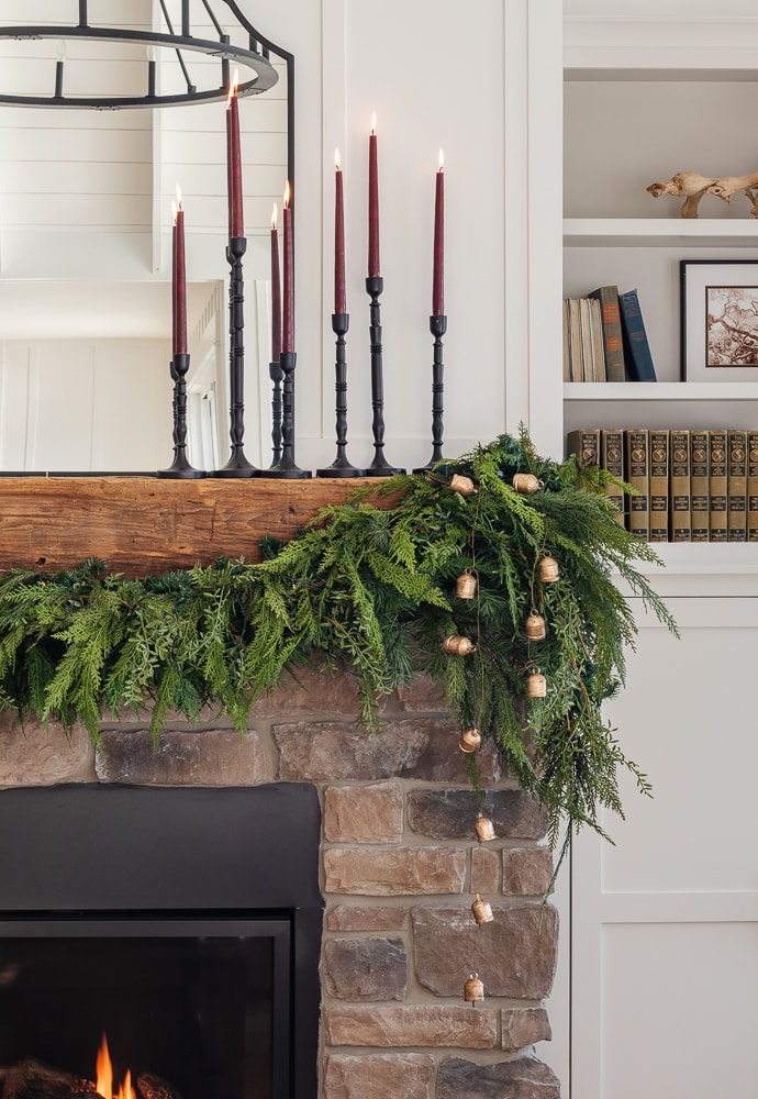 Christmas fireplace mantel decorated with fresh greenery, gold bells, and burgundy taper candles.