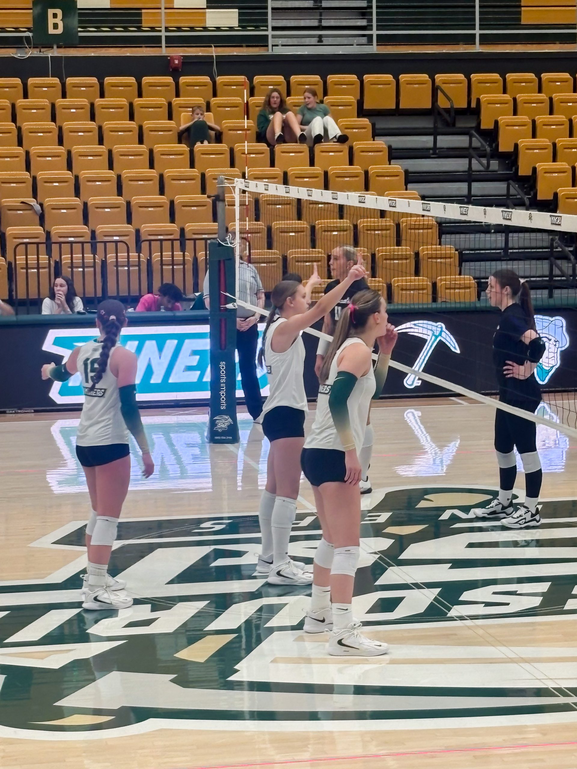 Missouri S&T women's volleyball players in white and green uniforms gather on the court during a home match at Gibson Arena.