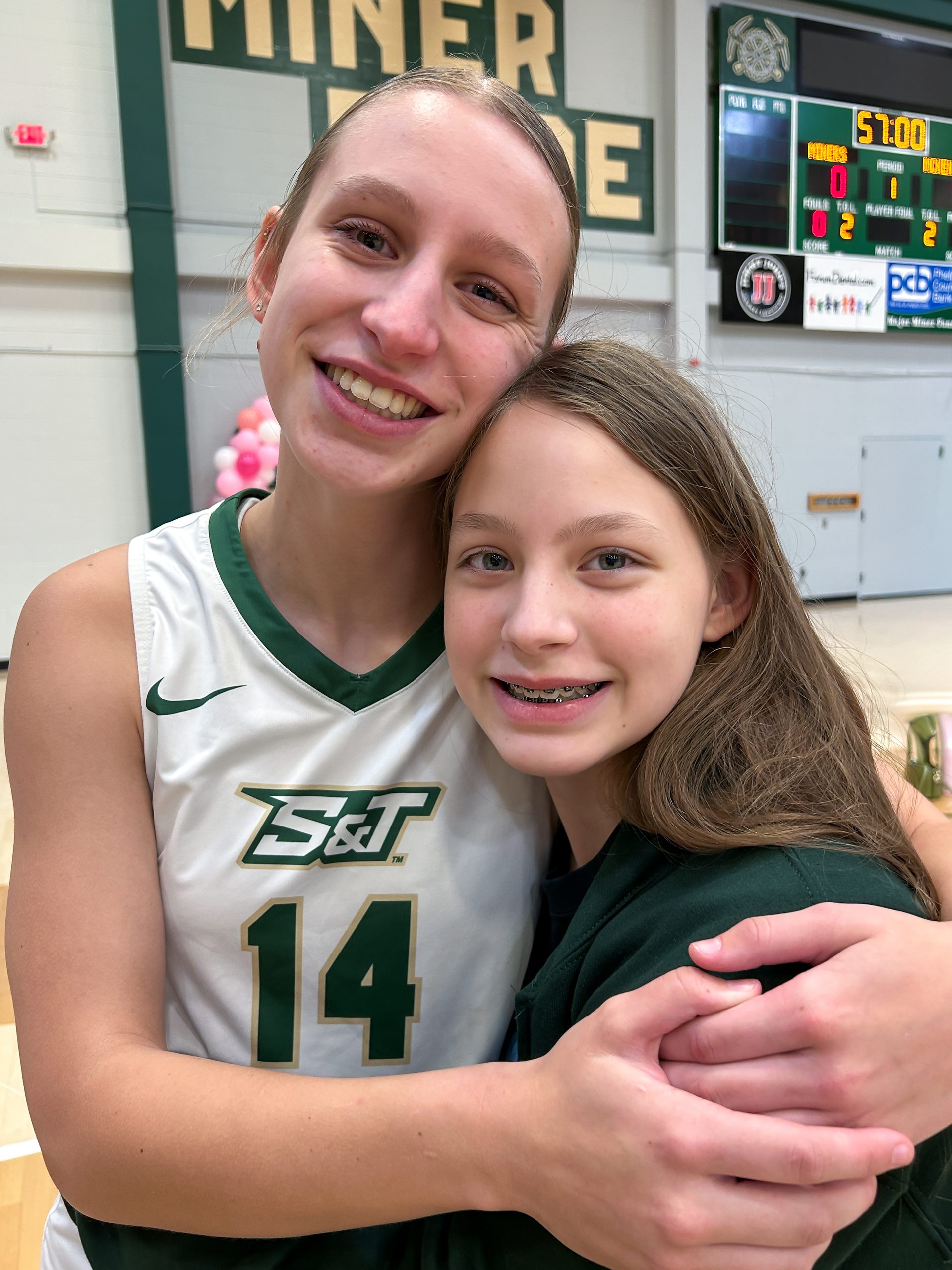 Two sisters smile and hug after a Missouri S&T volleyball game, with the older sister wearing her number 14 jersey